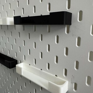 May include: A white pegboard with a series of black and white shelves attached. The shelves are rectangular and designed to fit into the pegboard holes. The shelves are in various sizes and orientations. The pegboard has a series of oval holes.