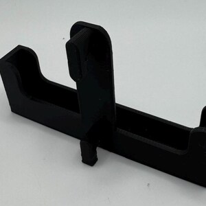 May include: A black plastic 3D printed phone stand with a slot for a phone and a raised section for a charging cable.