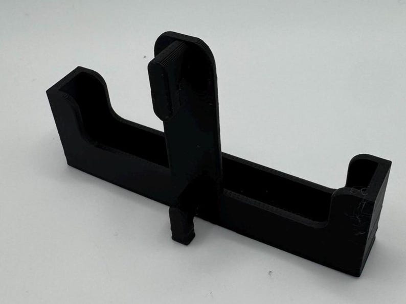 May include: A black plastic 3D printed phone stand with a slot for a phone and a raised section for a charging cable.