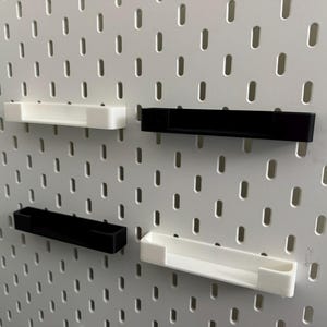 May include: Four small, rectangular shelves in black and white are mounted on a white pegboard. The shelves are designed to hold small items. The pegboard has evenly spaced holes. The shelves are made of plastic.