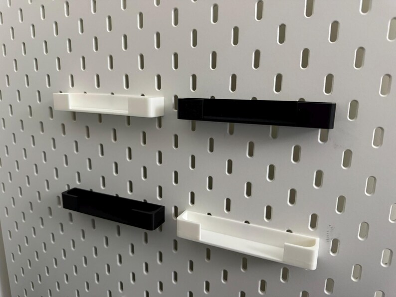 May include: Four small, rectangular shelves in black and white are mounted on a white pegboard. The shelves are designed to hold small items. The pegboard has evenly spaced holes. The shelves are made of plastic.