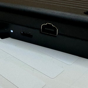 May include: A black rectangular electronic device with a USB port and a micro USB port on the side. The device is sitting on a white surface.