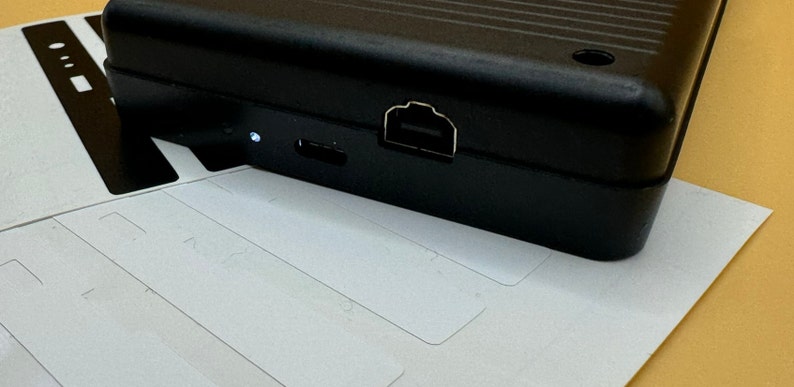 May include: A black rectangular electronic device with a USB port and a micro USB port on the side. The device is sitting on a white surface.