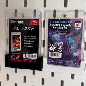Puede incluir: Dos protectores de tarjetas de pl&aacute;stico transparente, uno negro y otro blanco, est&aacute;n montados en una pared perforada blanca. El soporte izquierdo est&aacute; etiquetado como "Ultra PRO ONE-TOUCH" y el soporte derecho como "TITANSHIELD One-Click Magnetic Card Holders". Ambos soportes est&aacute;n dise&ntilde;ados para tarjetas de 35pt.