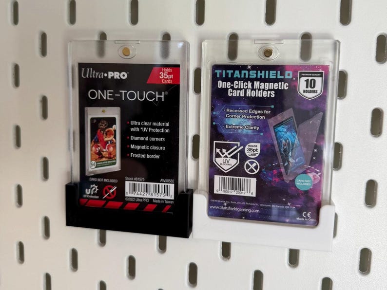 Puede incluir: Dos protectores de tarjetas de pl&aacute;stico transparente, uno negro y otro blanco, est&aacute;n montados en una pared perforada blanca. El soporte izquierdo est&aacute; etiquetado como "Ultra PRO ONE-TOUCH" y el soporte derecho como "TITANSHIELD One-Click Magnetic Card Holders". Ambos soportes est&aacute;n dise&ntilde;ados para tarjetas de 35pt.