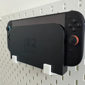 Puede incluir: Una consola y un mando Nintendo Switch negros están montados en una placa perforada blanca. El mando tiene botones negros con detalles en naranja y azul. La consola se sujeta con un soporte negro con soportes blancos.