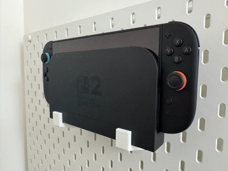 Op de afbeelding: Een zwarte Nintendo Switch-console en controller zijn op een witte pegboard gemonteerd. De controller heeft zwarte knoppen met oranje en blauwe accenten. De console wordt op zijn plaats gehouden door een zwarte houder met witte steunen.