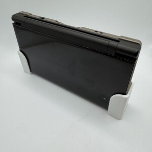 May include: A black laptop computer with a silver hinge is propped up by two white supports. The laptop is closed and has a glossy finish. The supports are rectangular and appear to be made of plastic.