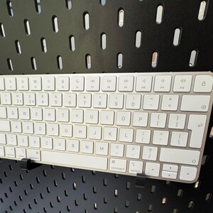 May include: A silver and white wireless keyboard with a full QWERTY layout. The keyboard is mounted on a black pegboard. The keys are white with black lettering. The keyboard has a command and option key.