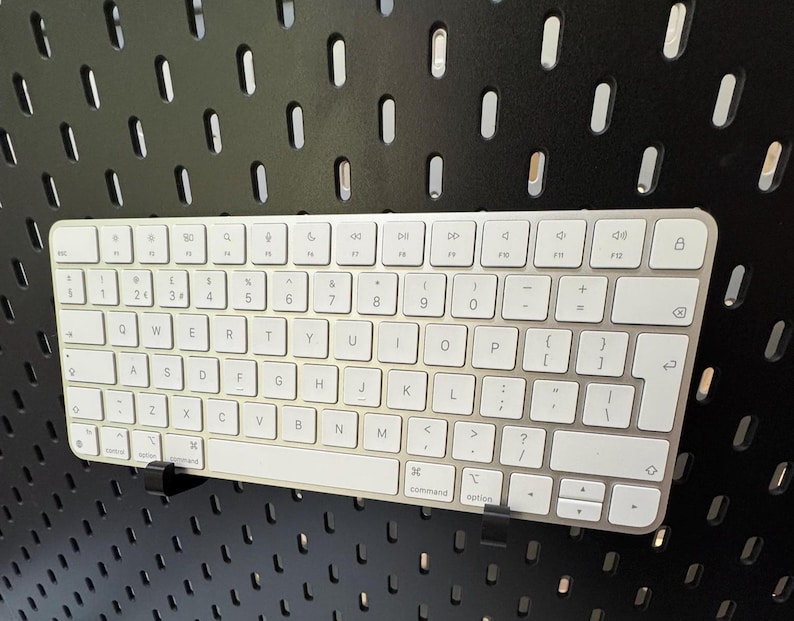 May include: A silver and white wireless keyboard with a full QWERTY layout. The keyboard is mounted on a black pegboard. The keys are white with black lettering. The keyboard has a command and option key.