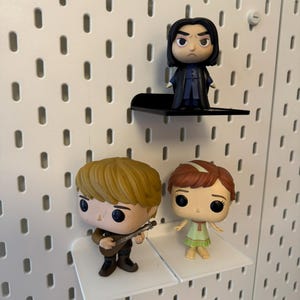 Pode incluir: Tr&ecirc;s figuras Pop Funko expostas em prateleiras de placa de pinos brancas. As figuras s&atilde;o uma figura masculina loira tocando uma guitarra, uma figura feminina ruiva vestindo um vestido verde e uma figura masculina de cabelo preto vestindo uma t&uacute;nica preta.