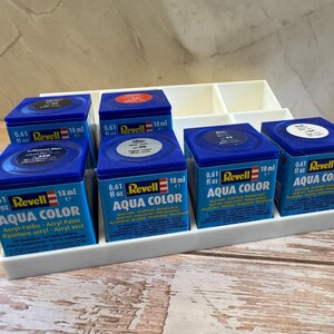 Peut inclure: Plusieurs pots de peinture acrylique Revell Aqua Color bleus, chacun contenant 18 ml de peinture. Les bo&icirc;tes sont dispos&eacute;es dans un organisateur en plastique blanc, avec le nom de la marque et le type de peinture visibles.