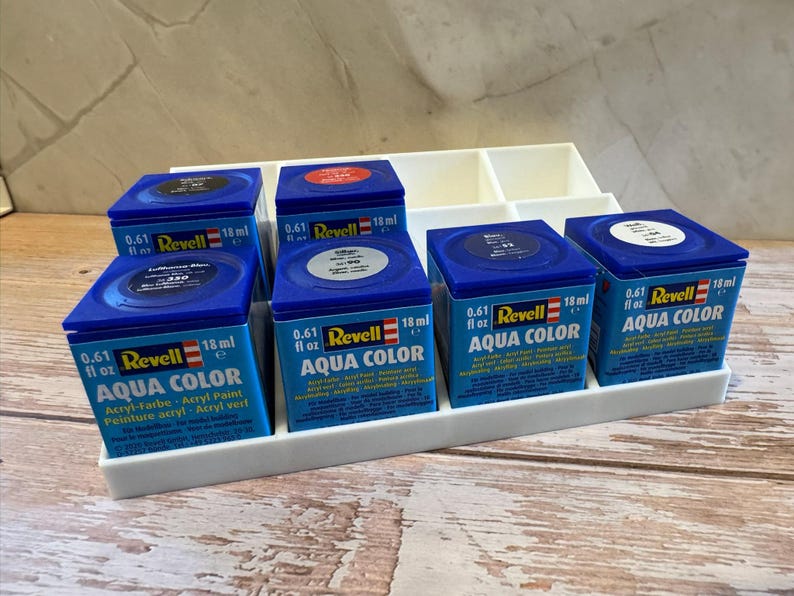 Peut inclure: Plusieurs pots de peinture acrylique Revell Aqua Color bleus, chacun contenant 18 ml de peinture. Les bo&icirc;tes sont dispos&eacute;es dans un organisateur en plastique blanc, avec le nom de la marque et le type de peinture visibles.