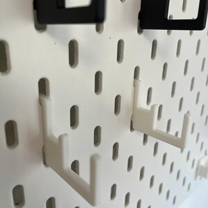 Puede incluir: Primer plano de un tablero perforado blanco con varios ganchos negros y blancos insertados. Los ganchos est&aacute;n dise&ntilde;ados para sujetar objetos y est&aacute;n hechos de pl&aacute;stico. El tablero perforado tiene agujeros ovalados espaciados.