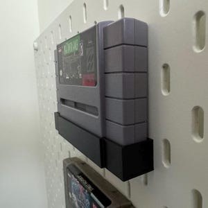 K&ouml;nnte beinhalten: Graue Super Nintendo-Spielkassette in einem schwarzen Halter, montiert an einer wei&szlig;en Lochwand. Das Spiel-Etikett ist sichtbar, mit gr&uuml;nen und roten Akzenten. Der Halter dient zur Pr&auml;sentation des Spiels.