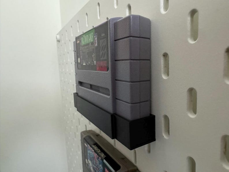 K&ouml;nnte beinhalten: Graue Super Nintendo-Spielkassette in einem schwarzen Halter, montiert an einer wei&szlig;en Lochwand. Das Spiel-Etikett ist sichtbar, mit gr&uuml;nen und roten Akzenten. Der Halter dient zur Pr&auml;sentation des Spiels.