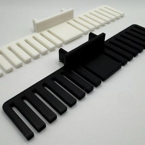 K&ouml;nnte beinhalten: Zwei 3D-gedruckte Kabelorganisatoren, einer schwarz und einer wei&szlig;. Jeder Organisator hat eine rechteckige Basis und mehrere Schlitze zum Halten von Kabeln.