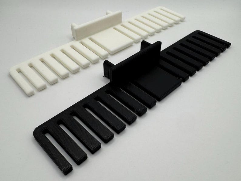 K&ouml;nnte beinhalten: Zwei 3D-gedruckte Kabelorganisatoren, einer schwarz und einer wei&szlig;. Jeder Organisator hat eine rechteckige Basis und mehrere Schlitze zum Halten von Kabeln.