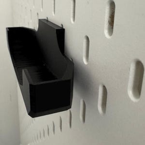 Op de afbeelding: Een zwarte, wandgemonteerde opbergorganisator van plastic met een rechthoekige vorm en een gebogen voorrand. De organisator is gemonteerd op een wit prikbord.