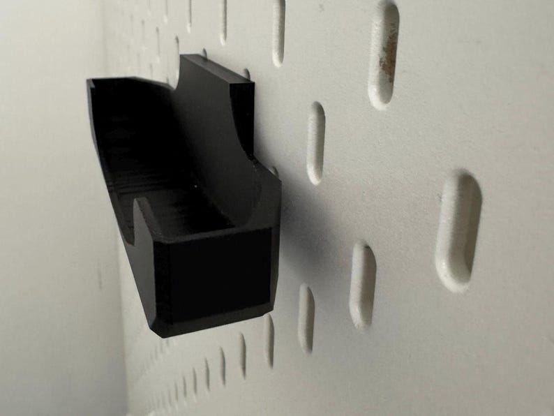Op de afbeelding: Een zwarte, wandgemonteerde opbergorganisator van plastic met een rechthoekige vorm en een gebogen voorrand. De organisator is gemonteerd op een wit prikbord.