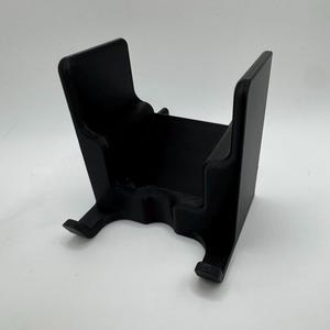 K&ouml;nnte beinhalten: Ein schwarzer, 3D-gedruckter St&auml;nder mit U-f&ouml;rmigem Design. Der St&auml;nder hat eine rechteckige Basis und zwei aufrechte St&uuml;tzen. Die St&uuml;tzen haben eine gebogene Oberseite und einen kleinen Vorsprung an der Unterseite. Der St&auml;nder ist zur Aufnahme eines Gegenstands bestimmt.