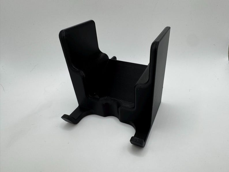 K&ouml;nnte beinhalten: Ein schwarzer, 3D-gedruckter St&auml;nder mit U-f&ouml;rmigem Design. Der St&auml;nder hat eine rechteckige Basis und zwei aufrechte St&uuml;tzen. Die St&uuml;tzen haben eine gebogene Oberseite und einen kleinen Vorsprung an der Unterseite. Der St&auml;nder ist zur Aufnahme eines Gegenstands bestimmt.