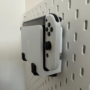 May include: A white Nintendo Switch console and controller are mounted on a white pegboard using black brackets. The controller has black buttons and joysticks. The pegboard is attached to a white wall.