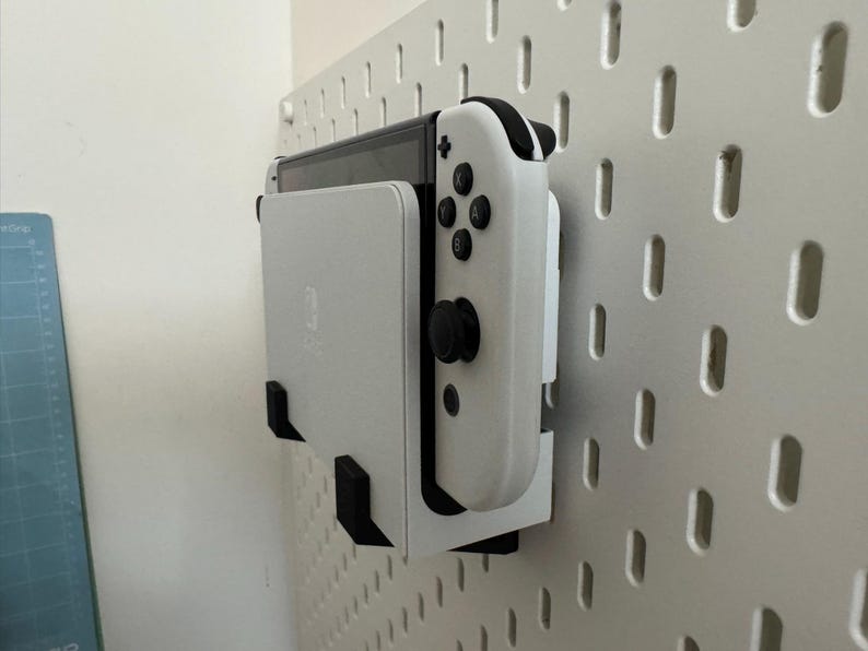 May include: A white Nintendo Switch console and controller are mounted on a white pegboard using black brackets. The controller has black buttons and joysticks. The pegboard is attached to a white wall.