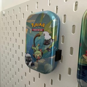 K&ouml;nnte beinhalten: Eine blau-gr&uuml;ne Pok&eacute;mon Trading Card Game-Box mit Zeichentrickfiguren und dem Pok&eacute;mon-Logo. Die Box ist an einer wei&szlig;en Lochwand befestigt.