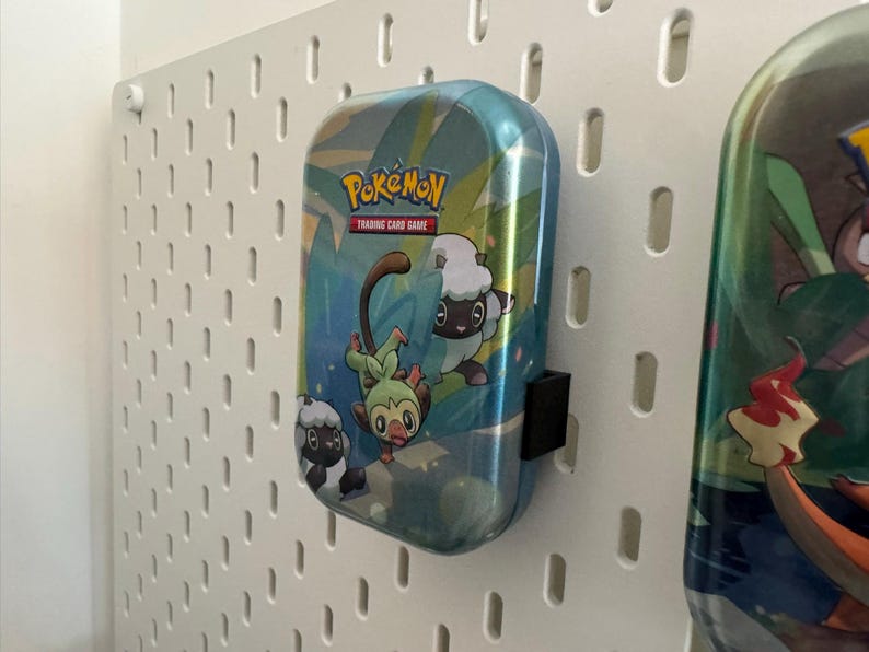 K&ouml;nnte beinhalten: Eine blau-gr&uuml;ne Pok&eacute;mon Trading Card Game-Box mit Zeichentrickfiguren und dem Pok&eacute;mon-Logo. Die Box ist an einer wei&szlig;en Lochwand befestigt.