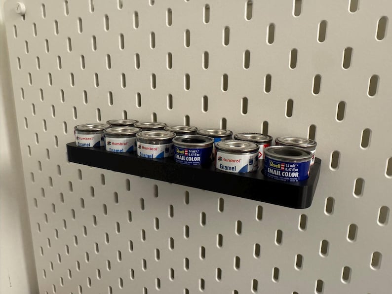May include: A black shelf holds several small paint cans against a white pegboard. The paint cans are silver with blue and red labels. The shelf is mounted on a wall with evenly spaced holes.
