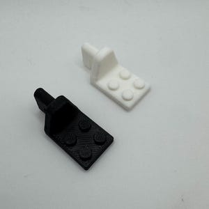 K&ouml;nnte beinhalten: Zwei kleine, 3D-gedruckte Kunststoffhalterungen. Eine ist schwarz, die andere wei&szlig;. Jede hat eine rechteckige Basis mit vier kreisf&ouml;rmigen Vorspr&uuml;ngen und einem vertikalen Pfosten. Die Halterungen sind wahrscheinlich f&uuml;r ein kleines Bauprojekt.