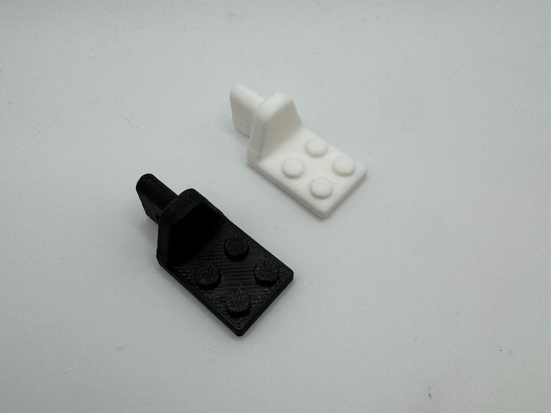 K&ouml;nnte beinhalten: Zwei kleine, 3D-gedruckte Kunststoffhalterungen. Eine ist schwarz, die andere wei&szlig;. Jede hat eine rechteckige Basis mit vier kreisf&ouml;rmigen Vorspr&uuml;ngen und einem vertikalen Pfosten. Die Halterungen sind wahrscheinlich f&uuml;r ein kleines Bauprojekt.