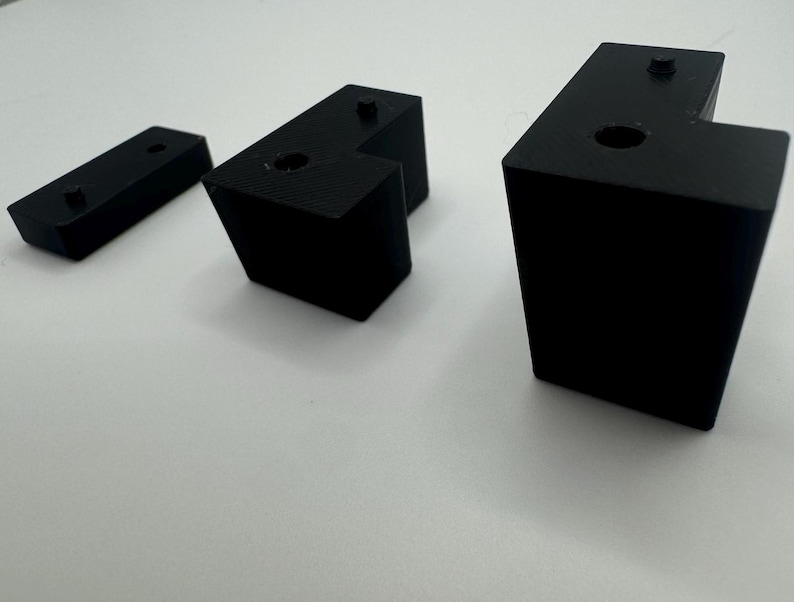 May include: Three black plastic corner brackets of different sizes. The brackets have a right angle shape with a hole in the centre of the top surface.