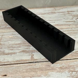 Puede incluir: Un organizador rectangular de madera negra con una serie de ranuras espaciadas uniformemente a lo largo del borde superior. El organizador est&aacute; sobre una superficie de madera clara y desgastada.