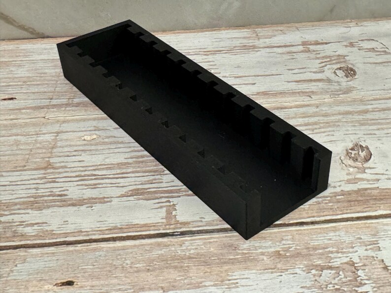 Puede incluir: Un organizador rectangular de madera negra con una serie de ranuras espaciadas uniformemente a lo largo del borde superior. El organizador est&aacute; sobre una superficie de madera clara y desgastada.
