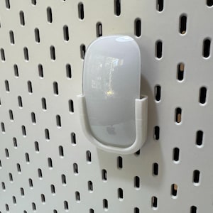 K&ouml;nnte beinhalten: Eine wei&szlig;e Apple Magic Mouse wird in einer wei&szlig;en, 3D-gedruckten Halterung gehalten, die an einer wei&szlig;en Lochplatte befestigt ist. Die Maus hat eine glatte, gl&auml;nzende Oberfl&auml;che und das Apple-Logo ist sichtbar. Die Lochplatte hat ein Gitter aus gleichm&auml;&szlig;ig verteilten, l&auml;nglichen L&ouml;chern.