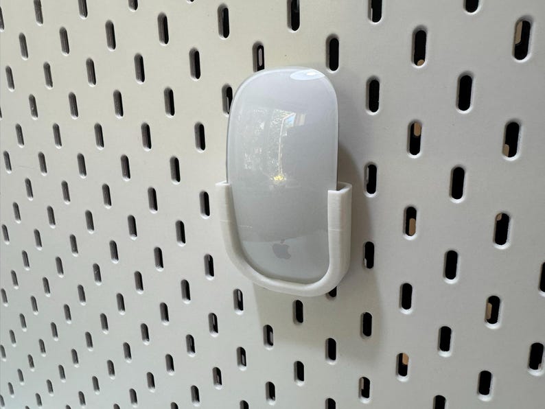 K&ouml;nnte beinhalten: Eine wei&szlig;e Apple Magic Mouse wird in einer wei&szlig;en, 3D-gedruckten Halterung gehalten, die an einer wei&szlig;en Lochplatte befestigt ist. Die Maus hat eine glatte, gl&auml;nzende Oberfl&auml;che und das Apple-Logo ist sichtbar. Die Lochplatte hat ein Gitter aus gleichm&auml;&szlig;ig verteilten, l&auml;nglichen L&ouml;chern.