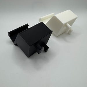 Op de afbeelding: Twee 3D-geprinte objecten. E&eacute;n is zwart, rechthoekig, met een sleuf en twee pennen. De andere is wit, kubusvormig, met een klein rechthoekig stuk en twee pennen. Beide staan op een witte achtergrond.