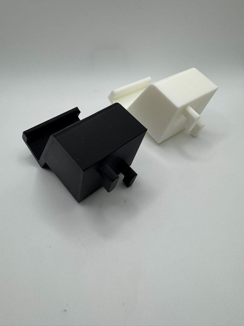Op de afbeelding: Twee 3D-geprinte objecten. E&eacute;n is zwart, rechthoekig, met een sleuf en twee pennen. De andere is wit, kubusvormig, met een klein rechthoekig stuk en twee pennen. Beide staan op een witte achtergrond.