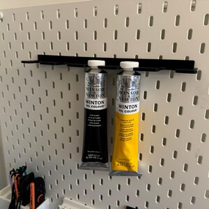 May include: Two tubes of Winsor & Newton Winton Oil Colour paint, one black and one yellow, are displayed on a black shelf attached to a white pegboard. The black tube is labeled "Lamp Black" and the yellow tube is labeled "Yellow Deep". Each tube is 6.75 US fl oz.
