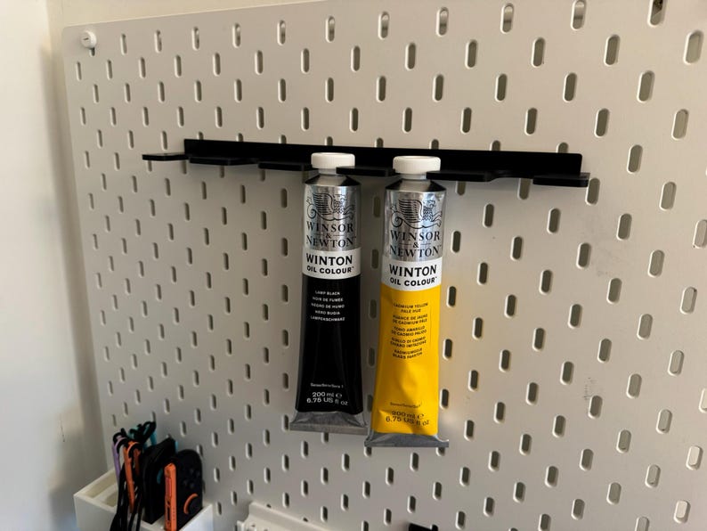 May include: Two tubes of Winsor & Newton Winton Oil Colour paint, one black and one yellow, are displayed on a black shelf attached to a white pegboard. The black tube is labeled "Lamp Black" and the yellow tube is labeled "Yellow Deep". Each tube is 6.75 US fl oz.