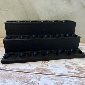 Puede incluir: Un organizador negro escalonado con tres niveles, cada uno con m&uacute;ltiples compartimentos circulares. El organizador est&aacute; hecho de un material mate y s&oacute;lido y descansa sobre una superficie de madera clara. El dise&ntilde;o es simple y funcional.