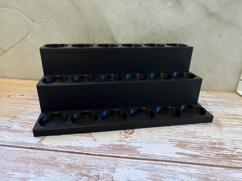Puede incluir: Un organizador negro escalonado con tres niveles, cada uno con m&uacute;ltiples compartimentos circulares. El organizador est&aacute; hecho de un material mate y s&oacute;lido y descansa sobre una superficie de madera clara. El dise&ntilde;o es simple y funcional.