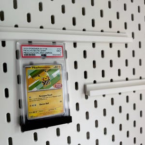 May include: A white pegboard with three white shelves. A graded Pokémon card featuring Pikachu is displayed on the pegboard. The card is in a protective case and is held in place by a black holder.