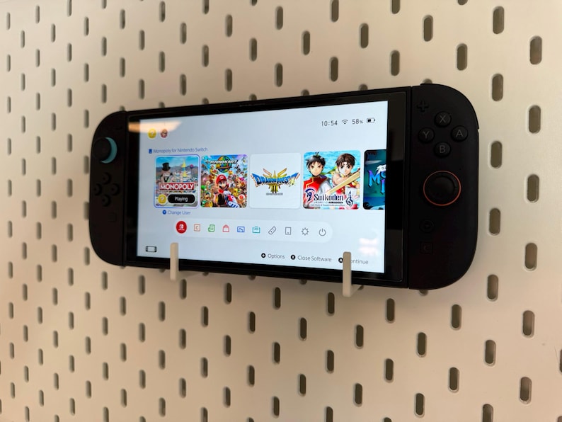 May include: A black handheld gaming console mounted on a white pegboard. The screen displays a game menu with several game icons. The console is held in place by white brackets.