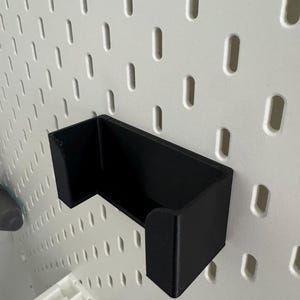 Op de afbeelding: Een zwarte, wandgemonteerde opbergorganisator van plastic met een rechthoekige vorm en een gebogen voorrand. De organisator is gemonteerd op een wit prikbord.
