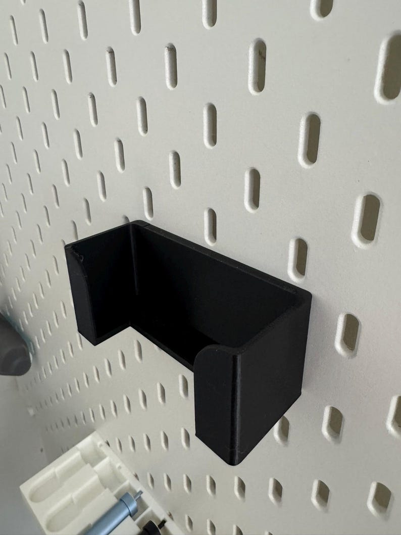 Op de afbeelding: Een zwarte, wandgemonteerde opbergorganisator van plastic met een rechthoekige vorm en een gebogen voorrand. De organisator is gemonteerd op een wit prikbord.