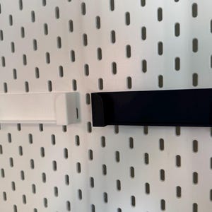 May include: Two shelf-like storage accessories, one white and one black, are mounted on a white pegboard. The shelves have a rectangular shape with curved edges, designed to hold small items. The pegboard has a pattern of evenly spaced oval holes.
