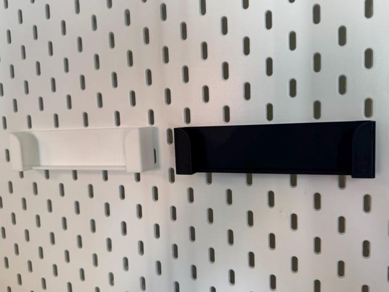May include: Two shelf-like storage accessories, one white and one black, are mounted on a white pegboard. The shelves have a rectangular shape with curved edges, designed to hold small items. The pegboard has a pattern of evenly spaced oval holes.